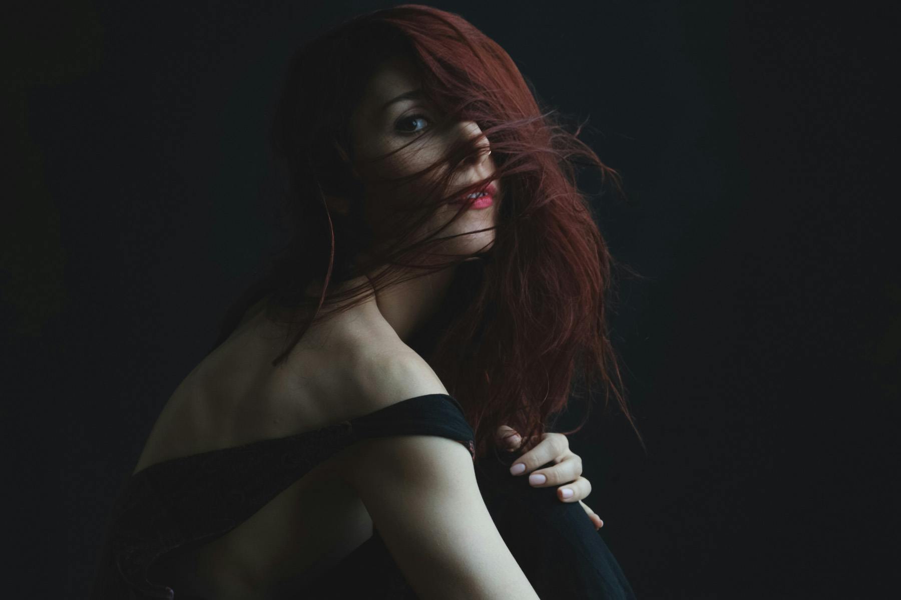 a portrait of Jana sitting in front of a black background with her dark red hair flowing in front of her face looking slightly towards the camera, her skin is light and you can clearly see the definition of her muscles.