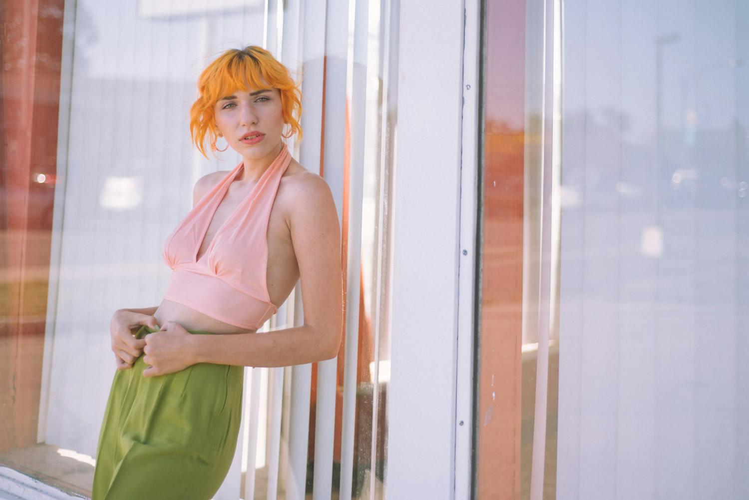 a portrait of Libby against a diner window holding her hands on the waistband of her pants, she is wearing a pink vintage top and green trousers and her hair is an orange color.
