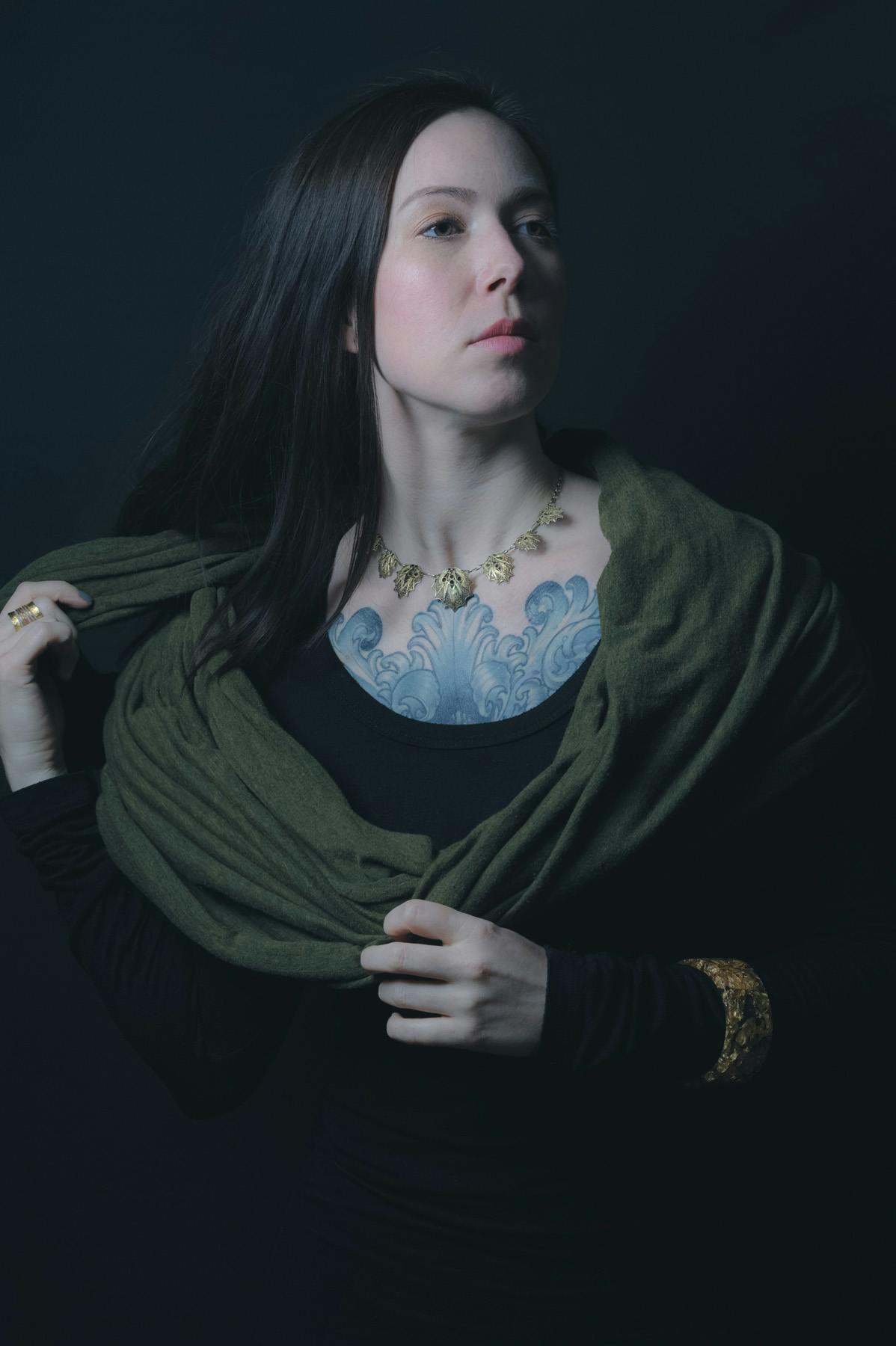 A portrait of Amelie wearing a black top with an ornate gold band around her left forearm, with a green scarf around her shoulder. she is in front of a black background.