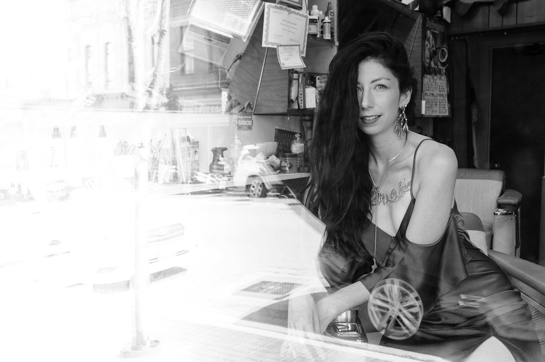 Black and white portrait of Jennifer sitting in a chair of a classic barber shop shot through a window