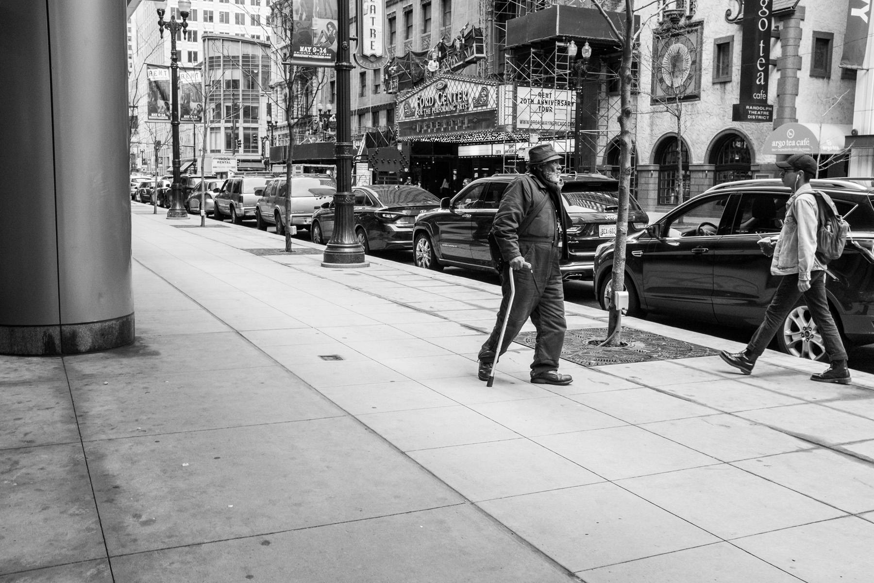 an older man bundled up walking through downtown Chicago with a cane.