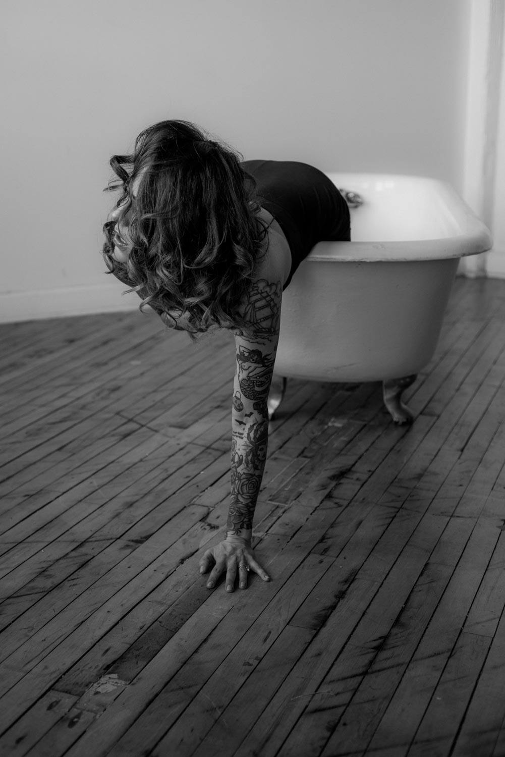 A black and white portrait of Christina crawling out of a claw foot bathtub sitting on a hardwood floor, she is holding herself up by a tattooed arm and is looking away from the camera with her hair over her face.
