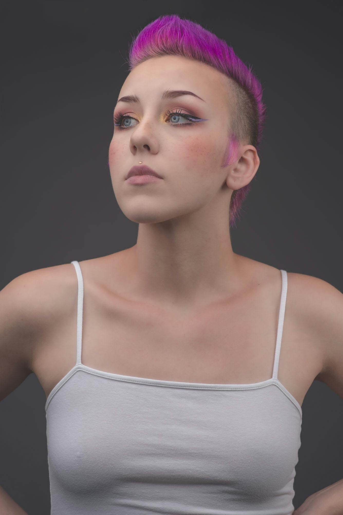 a portrait of Marybeth wearing a white tank top and a purple pink mohawk styled hairdo.