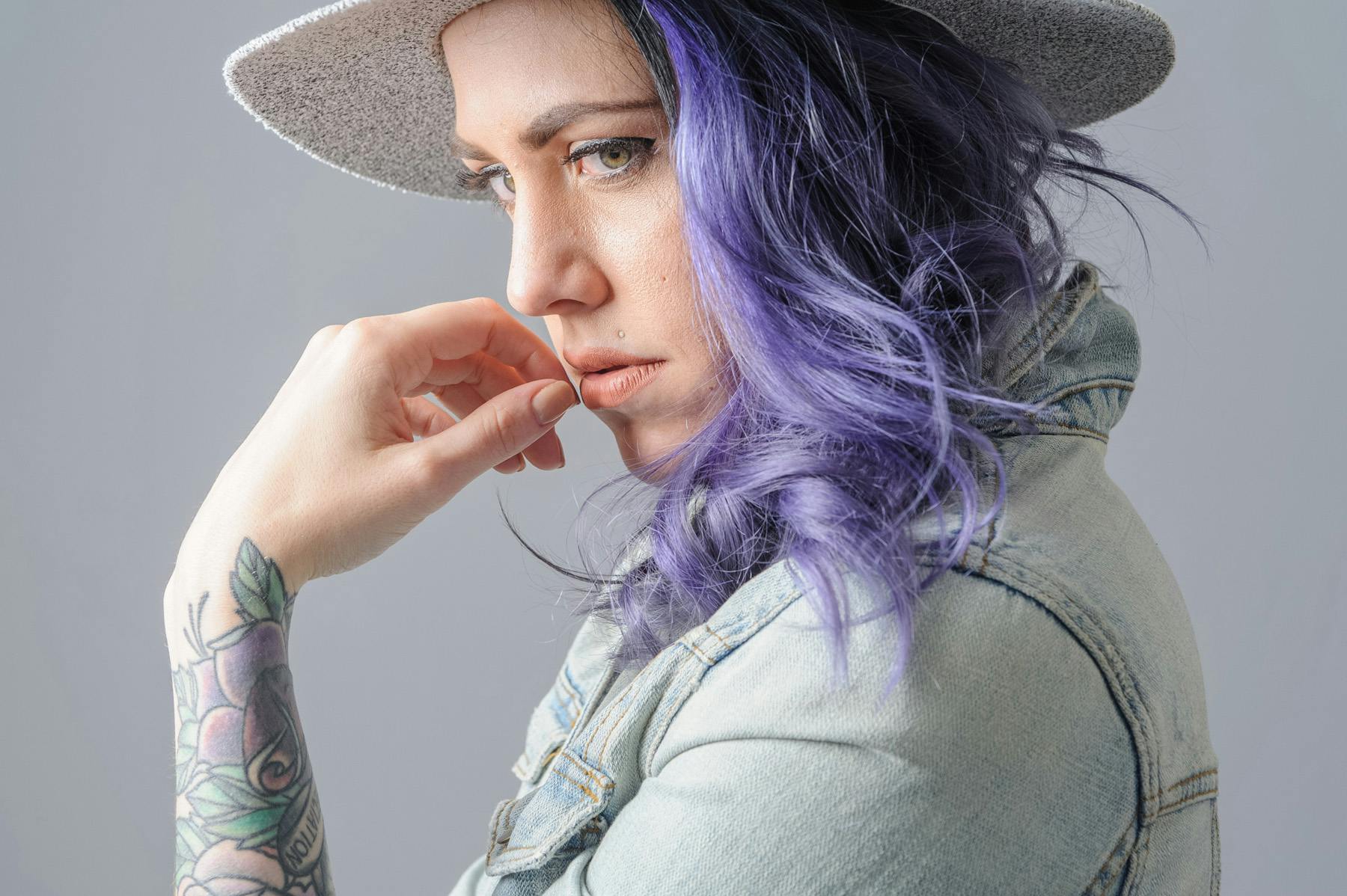  a portrait of Christina with purple hair and wearing a wide brim grey hat and faded blue jean jacket.