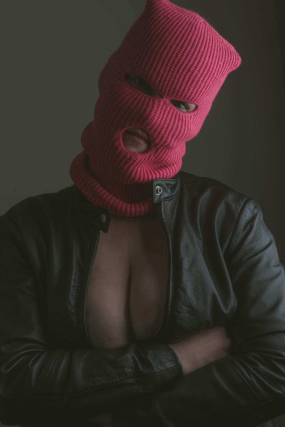a portrait of a model wearing only a black leather jacket and a pink ski mask crossing her arms under her chest.