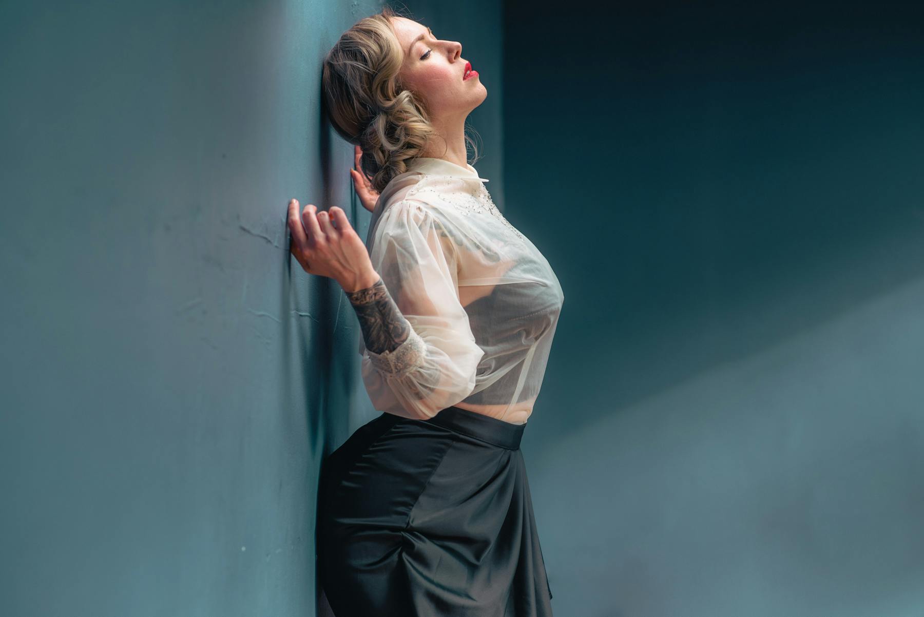 a portrait of Amelie wearing a white sheer blouse with a black bra beneath and a black skirt. she is leaning against a wall and is in profile.