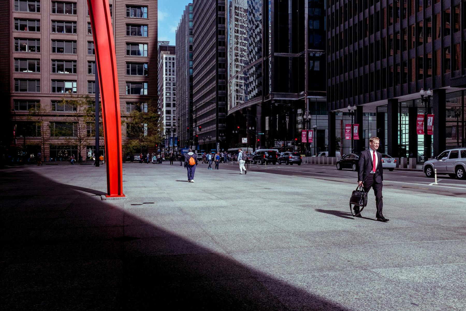 a photo of a man in a suit with a red tie, he is walking through Federal plaza and you can just see to the left of frame one of the legs from Calder's Flamingo.