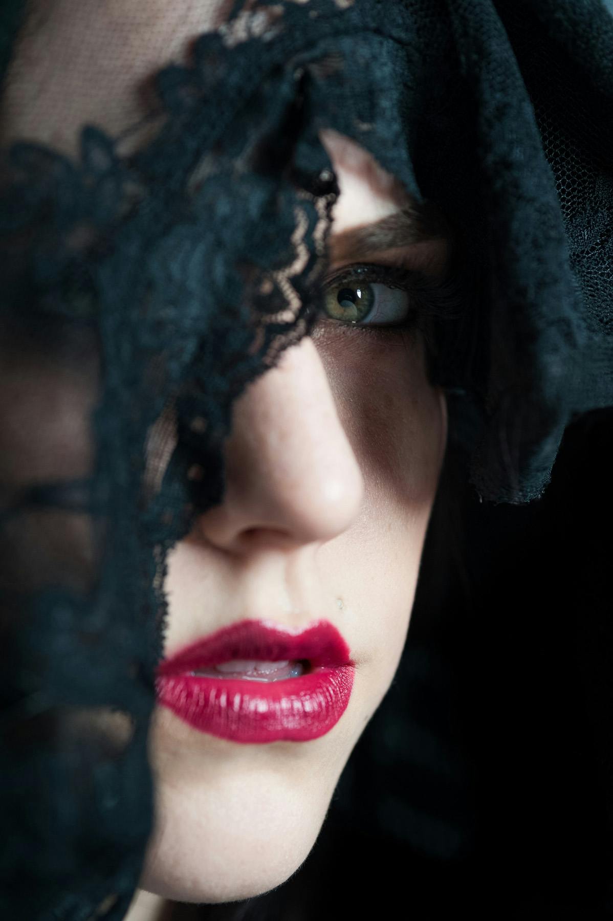 A close up color portrait of Christina wearing a black veil partially obscuring her face and you can clearly see her left eye a bright green color and her bright red lips set against her pale skin.