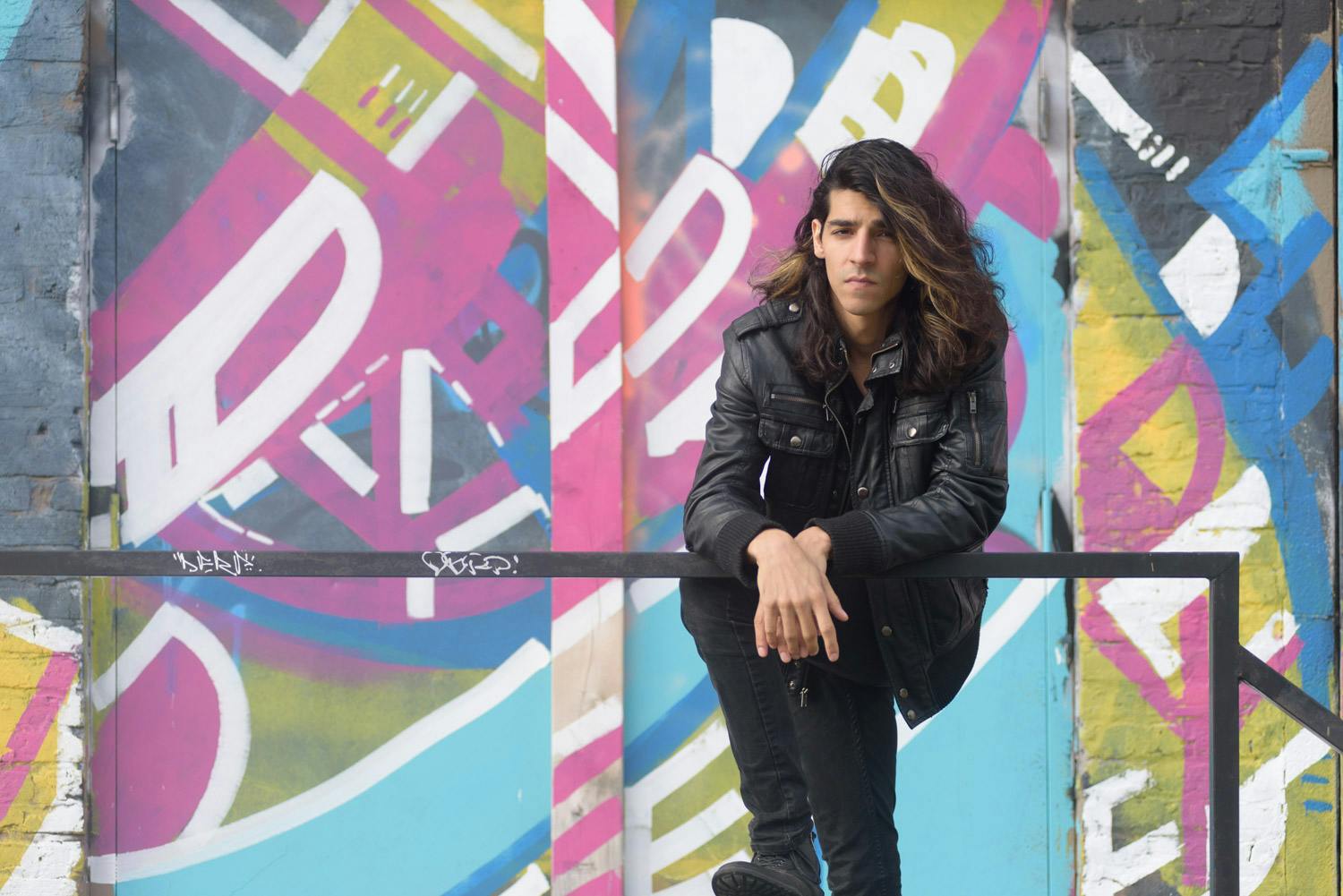 portrait of Matthew James Rao leaning against a railing towards the camera wearing a black leather jacket and pants in front of a pink, blue, yellow, and white graffiti wall