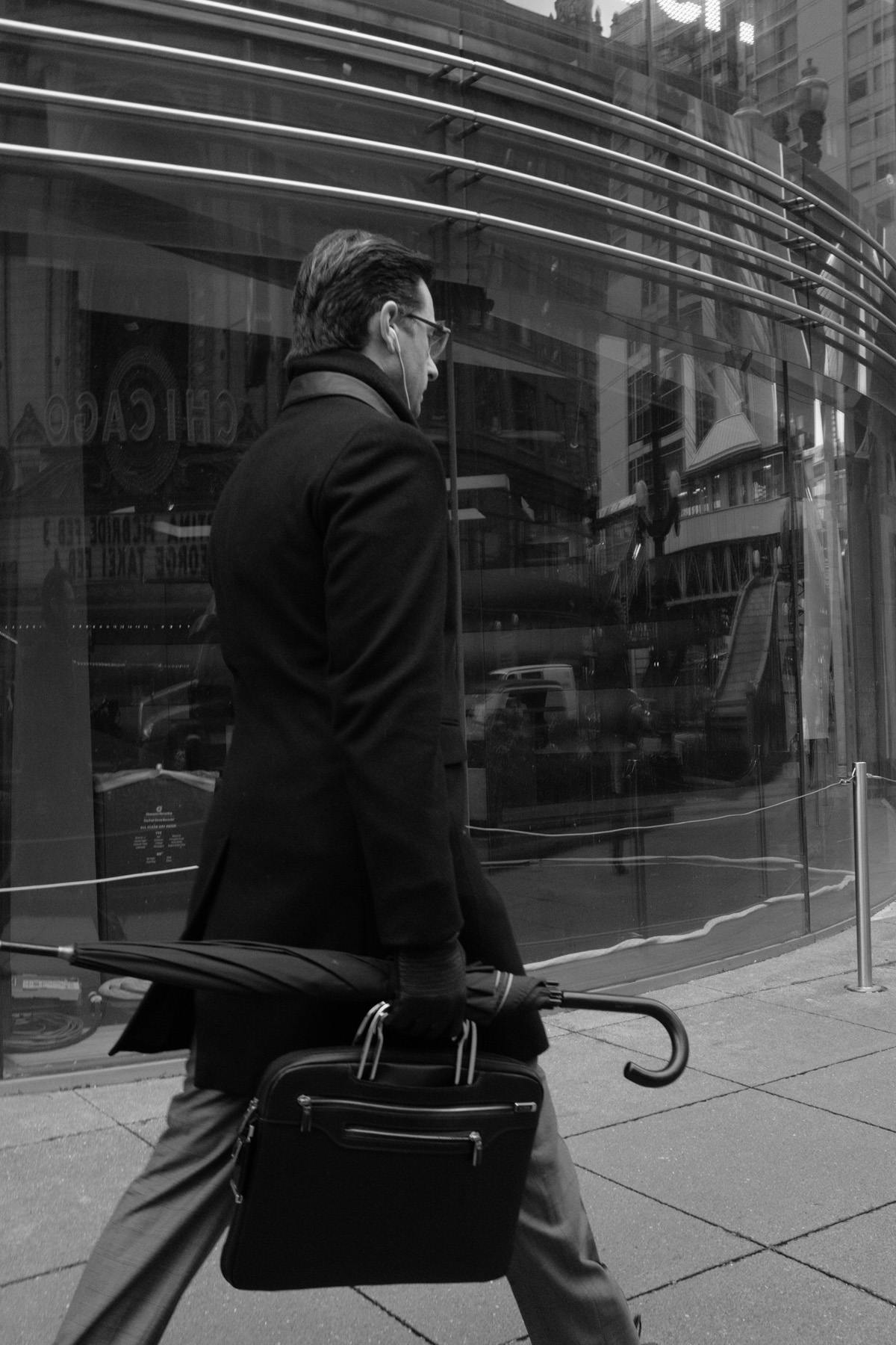 man wearing a black dress coat holding a briefcase and umbrella walking bristly to the right.