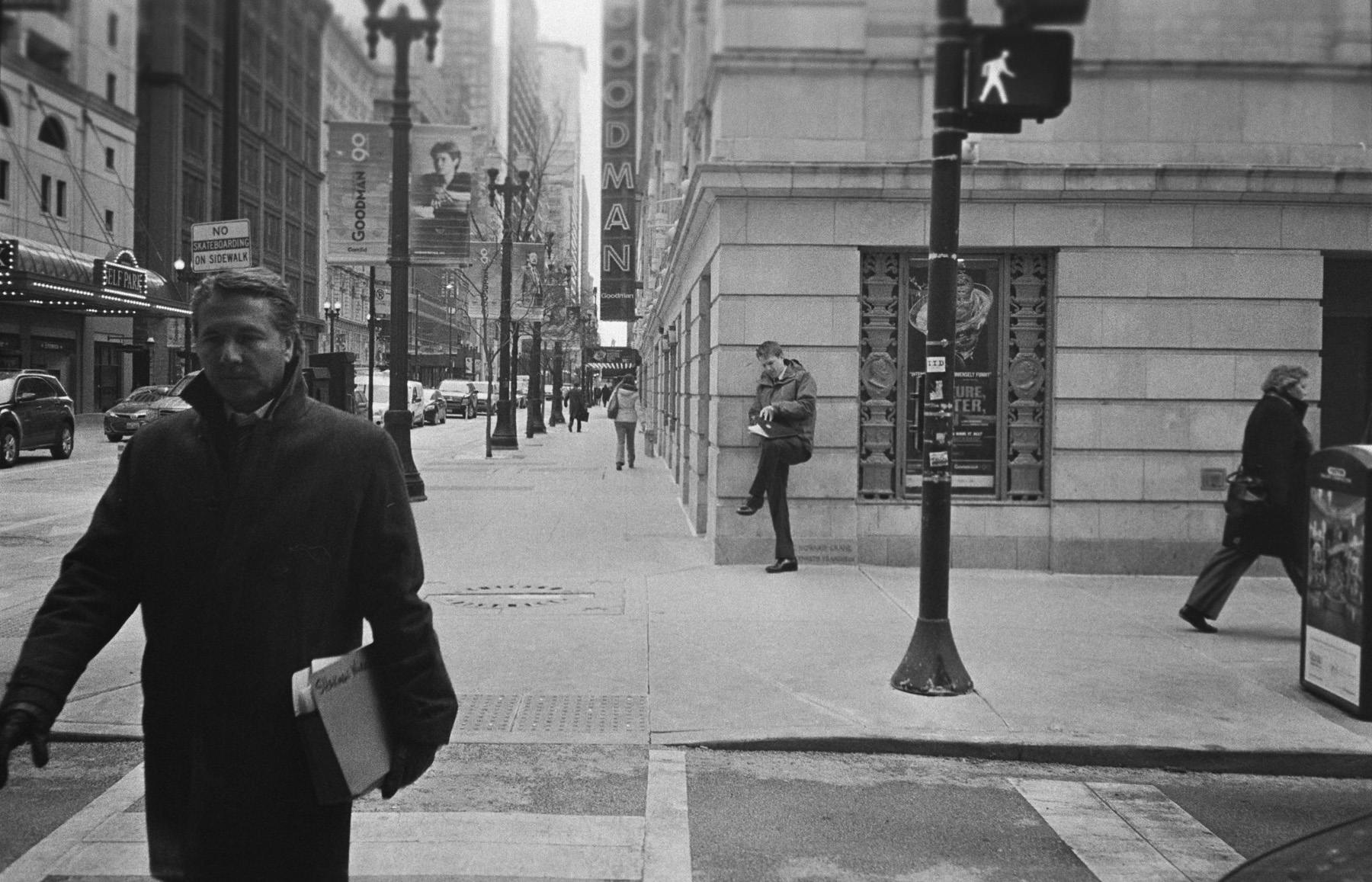 a film shot of a lawyer crossing the street in a dress winter coat, in the background you can see a man leaning up against a building looking through some documents on his knee.