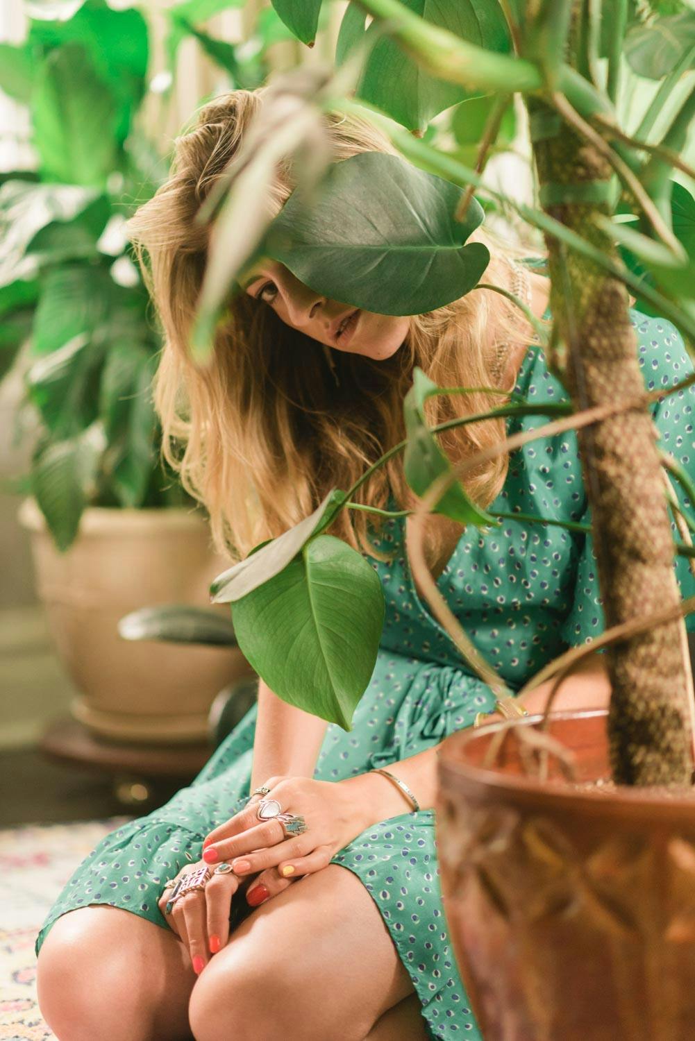 another portrait of Amanda with her plants slightly obscuring her face. she is wearing a green dress while kneeling on the floor.