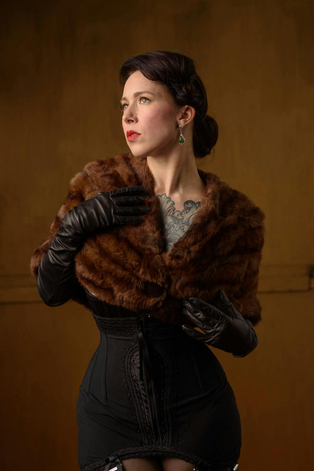 Portrait of woman wearing a fur shawl, long leather black gloves and an Edwardian styled corset in front of a copper colored backdrop.
